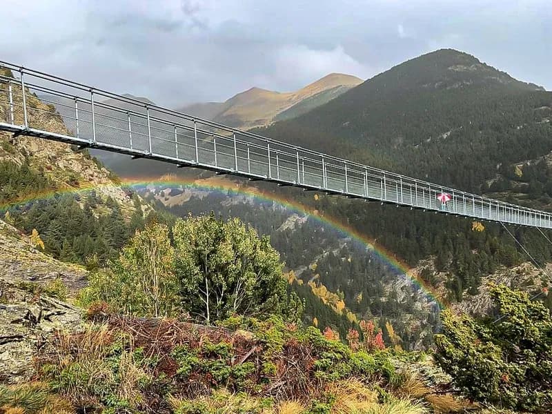 Un llarg pont penjant travessa una vall verda amb muntanyes al fons. Un arc de Sant Martí vibrant s'arqueja sota el pont, on una persona amb un paraigua vermell camina.