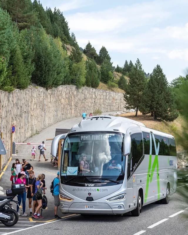 Un autobús turístico plateado con 'andorrabybus.com' y 'PONT TIBETA' en la pantalla está parado en una carretera, con varias personas de pie cerca de su parte delantera. Al fondo hay una ladera empinada cubierta de árboles con un muro de piedra.