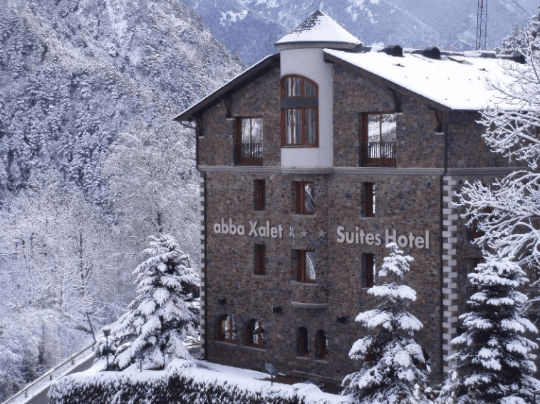 Un edificio de hotel de piedra, 'Abba Xalet Suites Hotel', se alza en medio de un paisaje montañoso cubierto de nieve con numerosos árboles nevados.