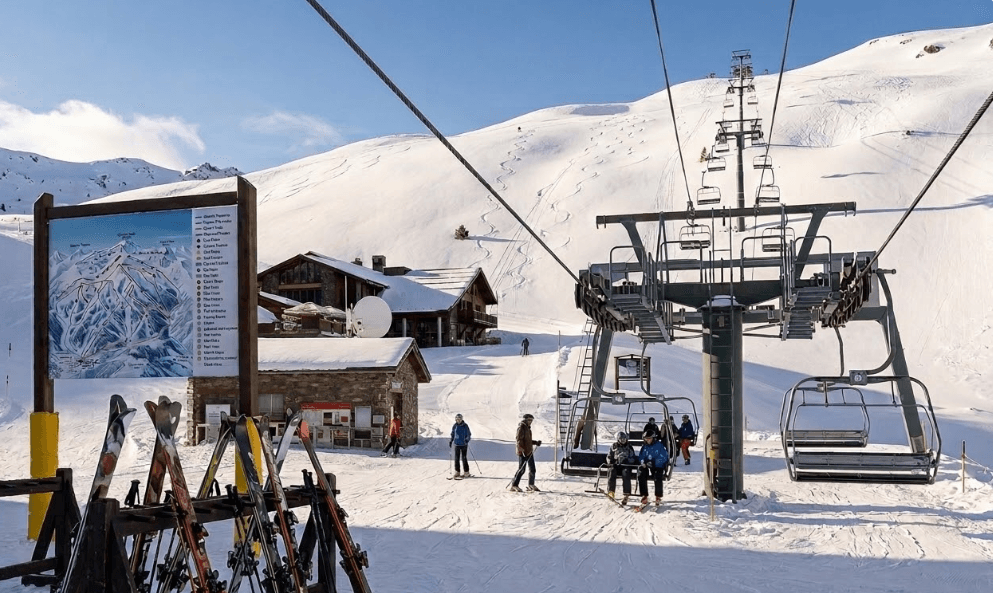 Una escena de estación de esquí con una estación de telesilla, esquiadores y un cartel con un mapa de pistas. Montañas cubiertas de nieve y chalets son visibles bajo un cielo azul claro.