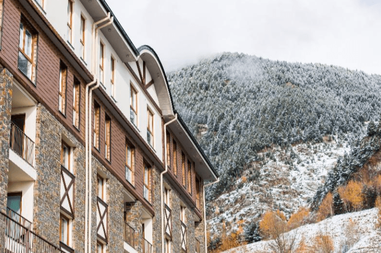 Un modern edifici d'apartaments amb façanes de pedra i fusta i balcons es troba davant d'una muntanya nevada amb pins i alguns arbres de fulles grogues.