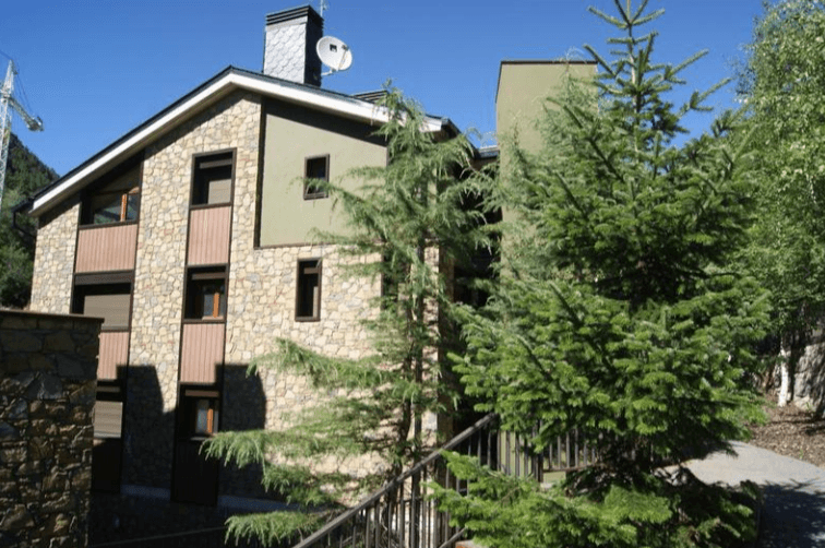 Un edificio moderno con revestimiento de piedra y paredes de estuco verde claro, con ventanas de marcos oscuros y detalles de madera, rodeado de frondosos pinos bajo un cielo azul claro.