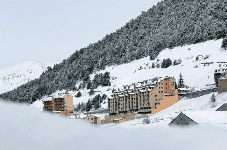 Edificios de apartamentos de varios pisos se asientan en una ladera de montaña nevada, parcialmente ocultos por una capa de niebla o nubes bajas. Pinos salpican el paisaje nevado.