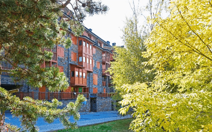 Un edificio de varios pisos con paredes de piedra y balcones de madera se encuentra entre árboles con follaje verde y amarillo brillante de otoño. Un camino pavimentado pasa frente al edificio.
