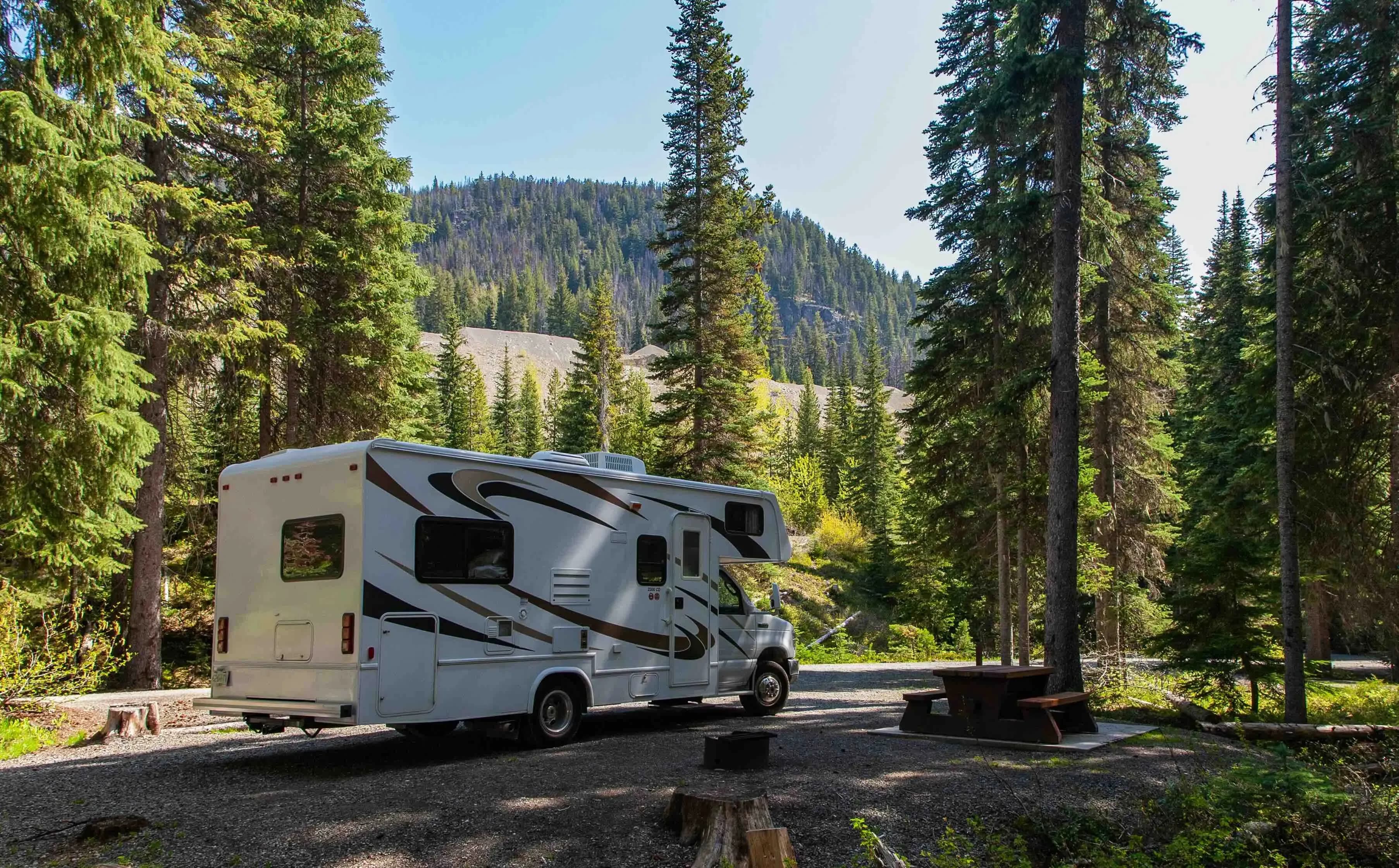 Una autocaravana blanca y marrón está estacionada en un campamento de grava rodeada de altos árboles de hoja perenne. Una mesa de picnic y una fogata son visibles a la derecha, con una montaña boscosa al fondo bajo un cielo despejado.