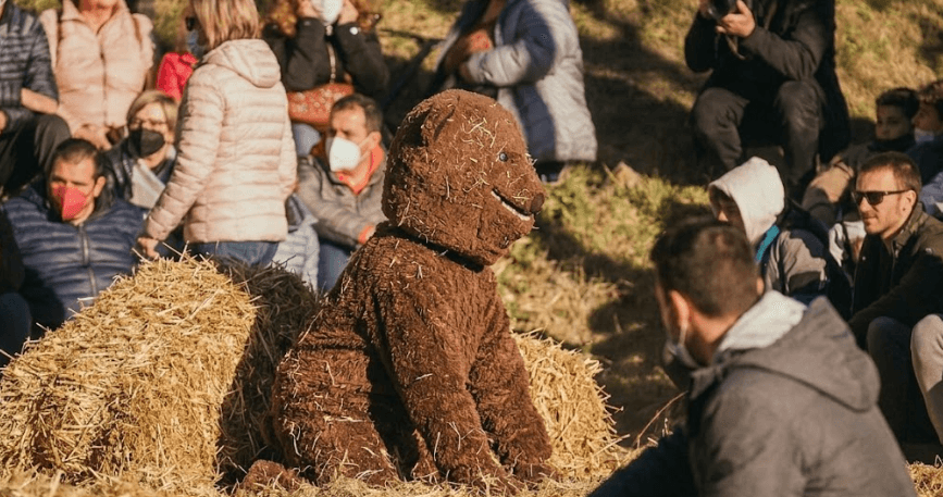 Une personne vêtue d'un costume d'ours brun hirsute est assise sur des bottes de foin, entourée d'une foule de spectateurs, certains portant des masques faciaux, lors d'un événement en plein air.