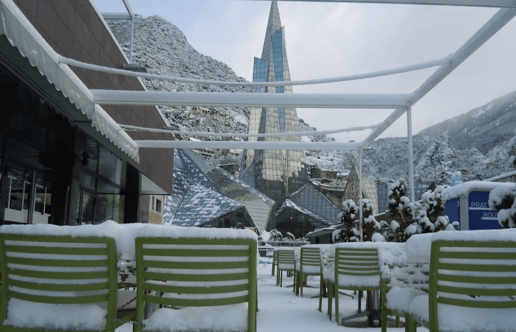 Una terrassa exterior coberta de neu amb cadires i taules verdes, amb vistes a un edifici modern de vidre i muntanyes nevades sota un cel ennuvolat.
