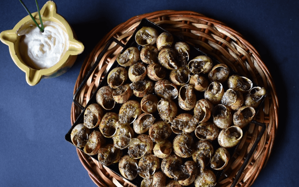 Un primer plano muestra caracoles cocidos en sus conchas sobre una bandeja de metal negro, apoyada en una cesta de mimbre, con un mortero amarillo de salsa blanca y cebollino a un lado.