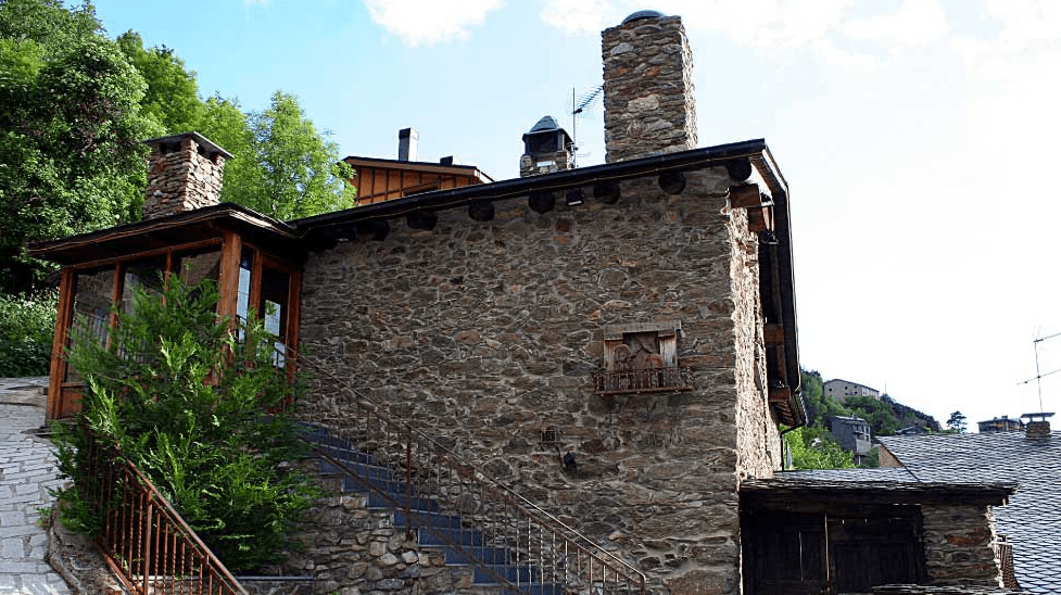 Un edificio tradicional de piedra con un porche de madera y una chimenea de piedra prominente bajo un cielo azul. Unas escaleras con barandilla de metal suben hacia la entrada, rodeadas de vegetación.