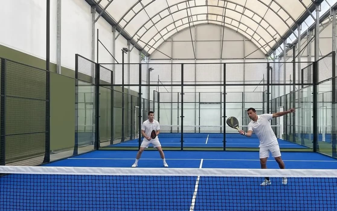 Dos homes amb roba esportiva blanca juguen a pàdel en una pista blava coberta amb una xarxa en primer pla. Un home està en posició d'espera, mentre que l'altre està a mig cop amb la seva pala.