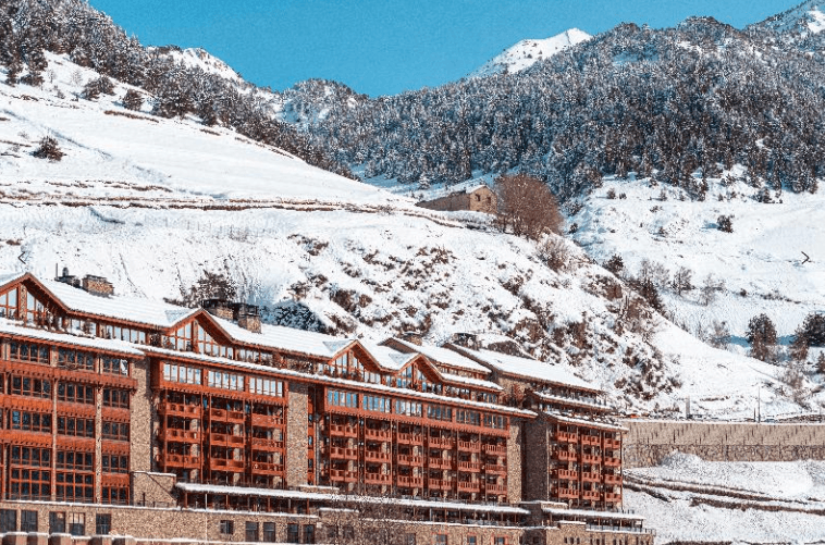 Un gran edificio moderno con balcones de madera y paredes de piedra se encuentra en un paisaje montañoso nevado bajo un cielo azul claro.