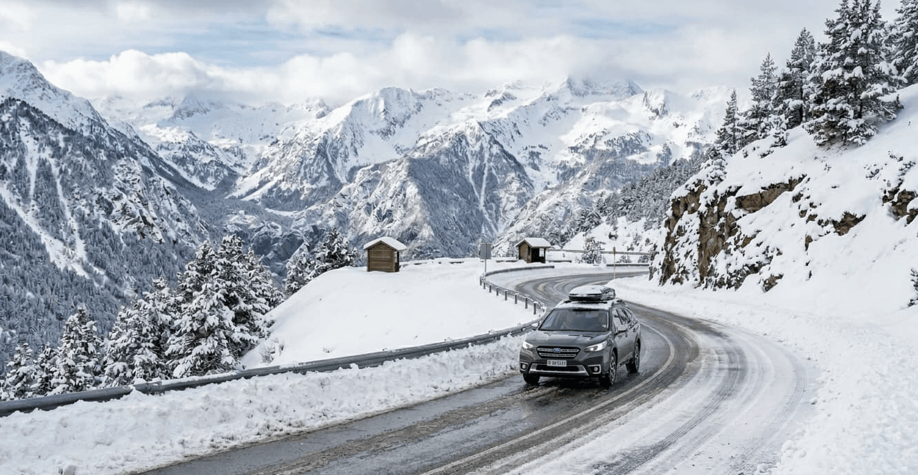 Un SUV gris oscuro circula por una carretera de montaña sinuosa y parcialmente cubierta de nieve, rodeada de picos nevados y pinos bajo un cielo nublado.