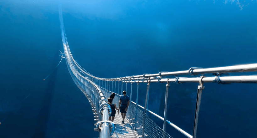 Dos personas caminan por un largo y estrecho puente colgante que atraviesa un profundo abismo azul.