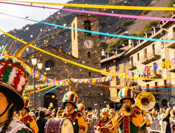 Un défilé de rue animé présente des musiciens costumés jouant d'instruments sous des confettis et des serpentins colorés, avec une église en pierre et des montagnes en arrière-plan.