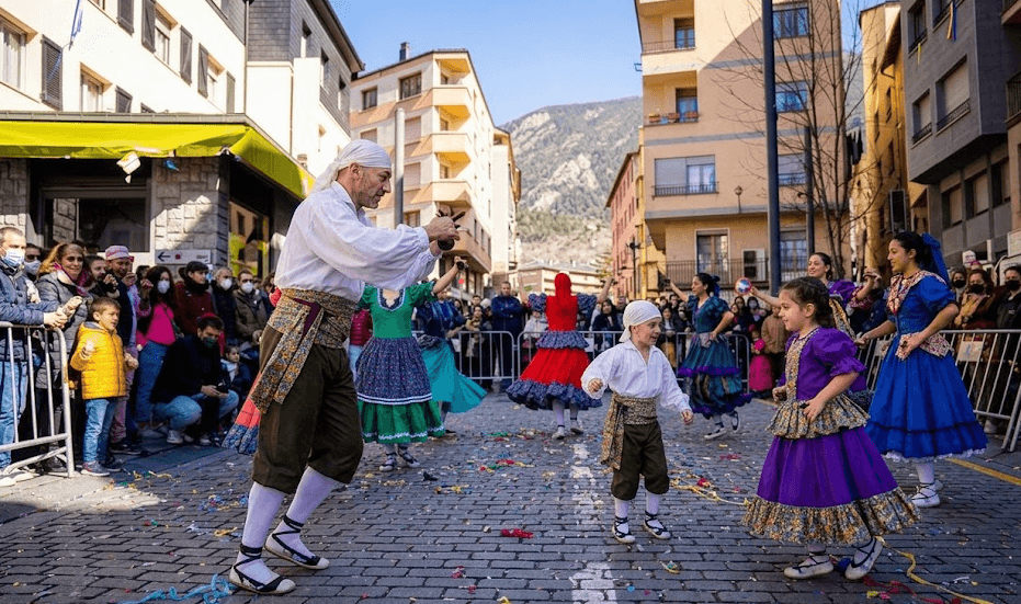 Des danseurs en costumes folkloriques traditionnels se produisent dans une rue ensoleillée jonchée de confettis, sous le regard des spectateurs. Un homme adulte et un jeune garçon en chemises blanches et écharpes dansent aux côtés de femmes et de filles en robes colorées à plusieurs couches.