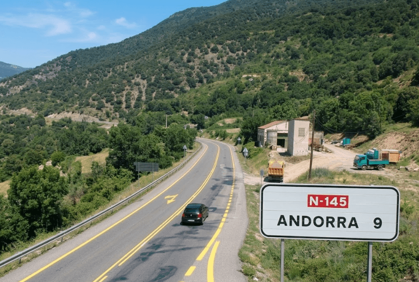 Un cotxe negre circula per una carretera de muntanya sinuosa amb un senyal que indica "N-145 ANDORRA 9" en primer pla. Al fons es veuen muntanyes verdes i alguns edificis.