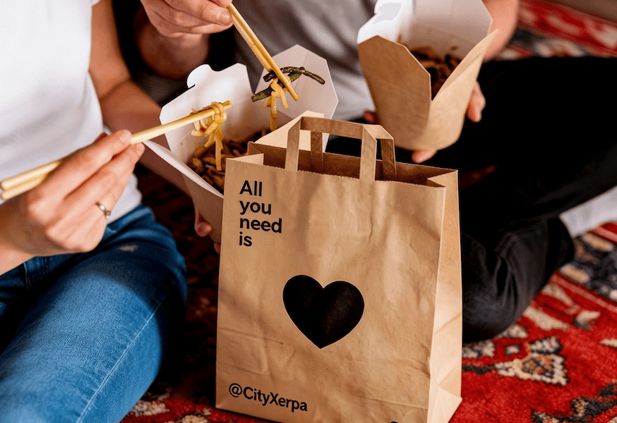 Dos personas comen fideos de cajas de cartón para llevar con palillos, sentadas sobre una alfombra estampada. Una bolsa de papel marrón con el texto "All you need is" y un logo de corazón está entre ellos.