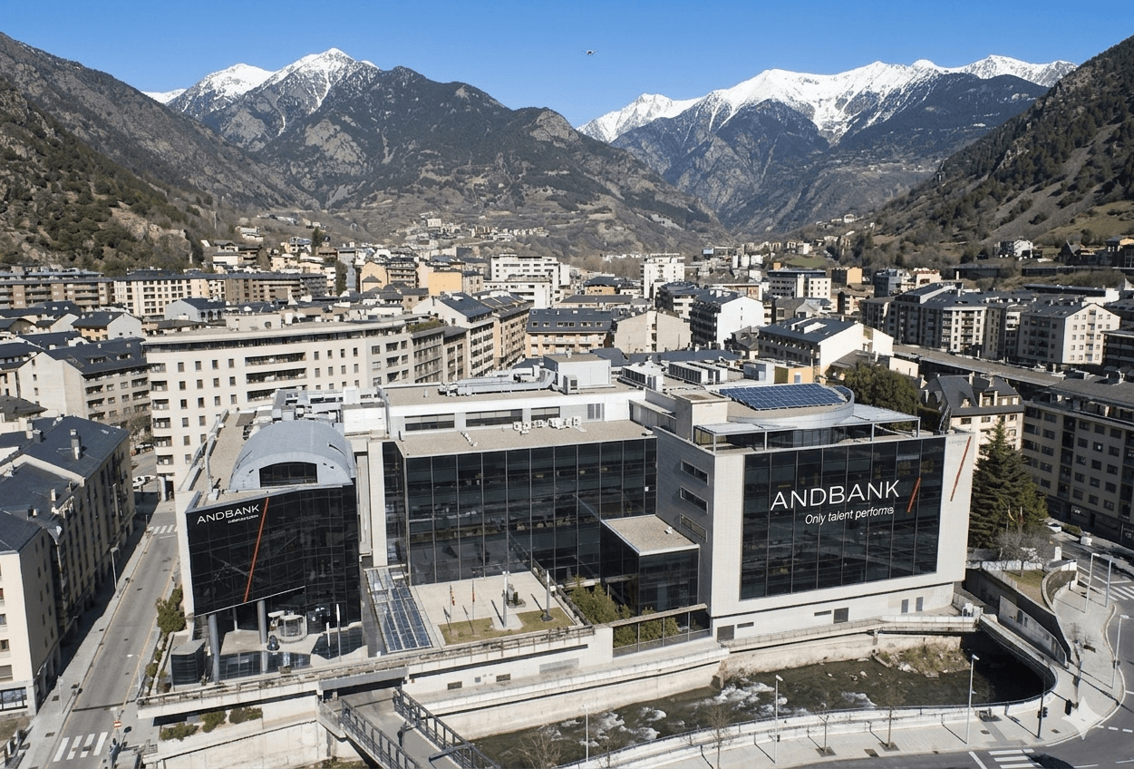 Vista aèria d'un modern edifici bancari amb façanes de vidre, situat al costat d'un riu en una vall urbana, envoltat d'edificis residencials i muntanyes nevades sota un cel clar.