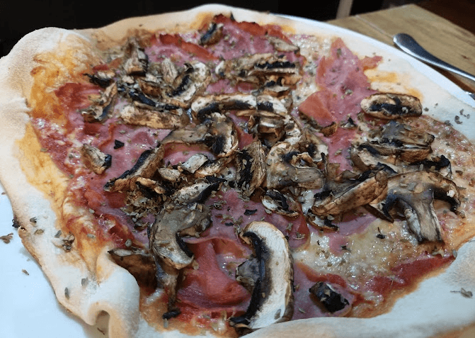 Vista de prop d'una pizza coberta amb salsa de tomàquet, formatge fos, talls de pernil i abundants bolets saltejats, espolvorada amb herbes seques. La massa és gruixuda i lleugerament daurada.