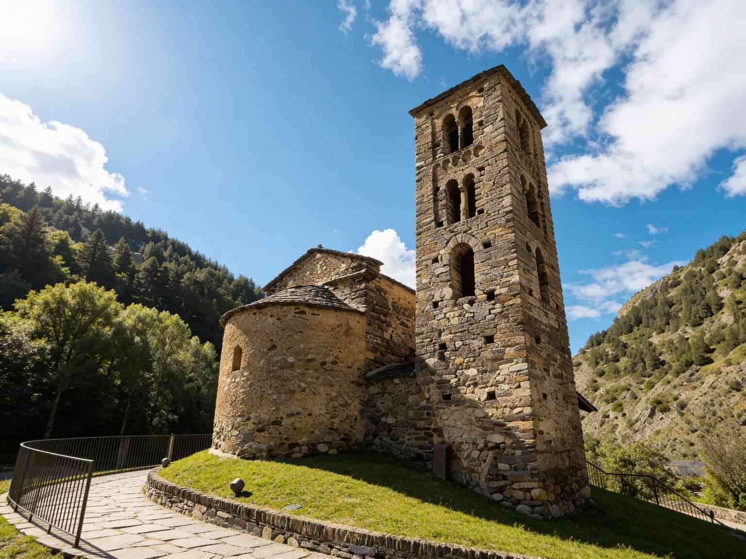 Una iglesia románica de piedra con un campanario alto y cuadrado se alza sobre una colina cubierta de hierba. Un camino de piedra con barandilla conduce a la iglesia, con montañas boscosas y un cielo azul de fondo.