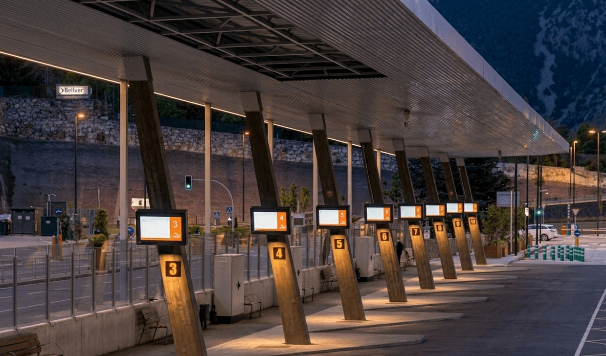 Una estació d'autobusos moderna i ben il·luminada al capvespre, amb múltiples andanes, pilars inclinats i pantalles digitals que mostren números. La llarga marquesina està il·luminada i es veuen muntanyes al fons.