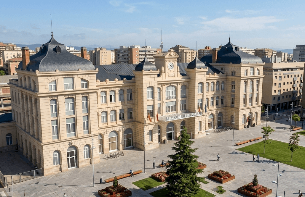 Un gran edifici de pedra de color clar amb arquitectura clàssica, teulades corbes fosques i un rellotge central, identificat com a "Estació de Lleida Pirineus", s'alça darrere d'una moderna plaça pública amb gespa verda, arbres i parterres sota un cel clar.