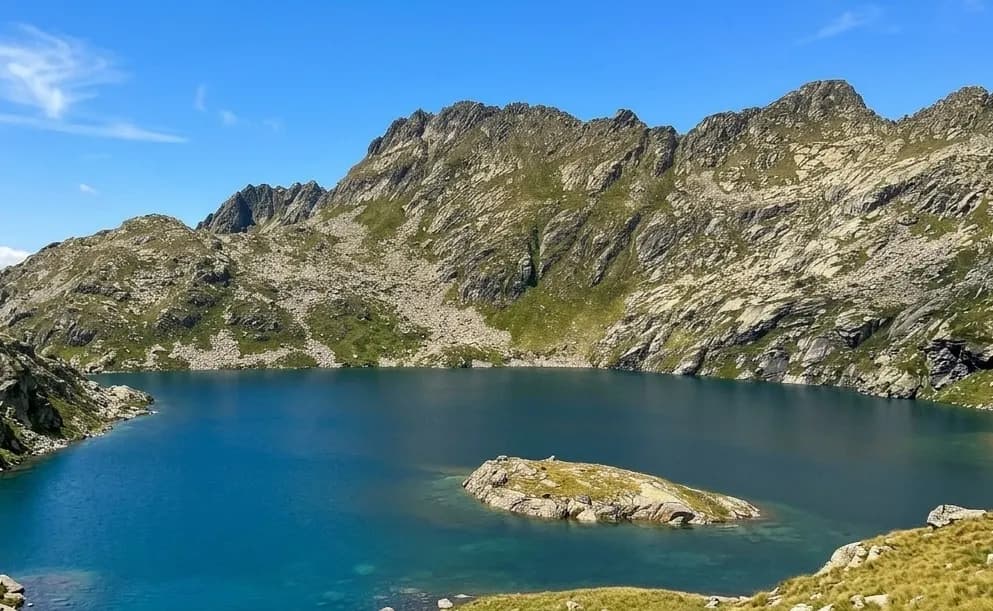 Un llac de muntanya de color blau clar envoltat de muntanyes rocoses i verdes sota un cel blau brillant. Una petita illa rocosa és visible al mig del llac.