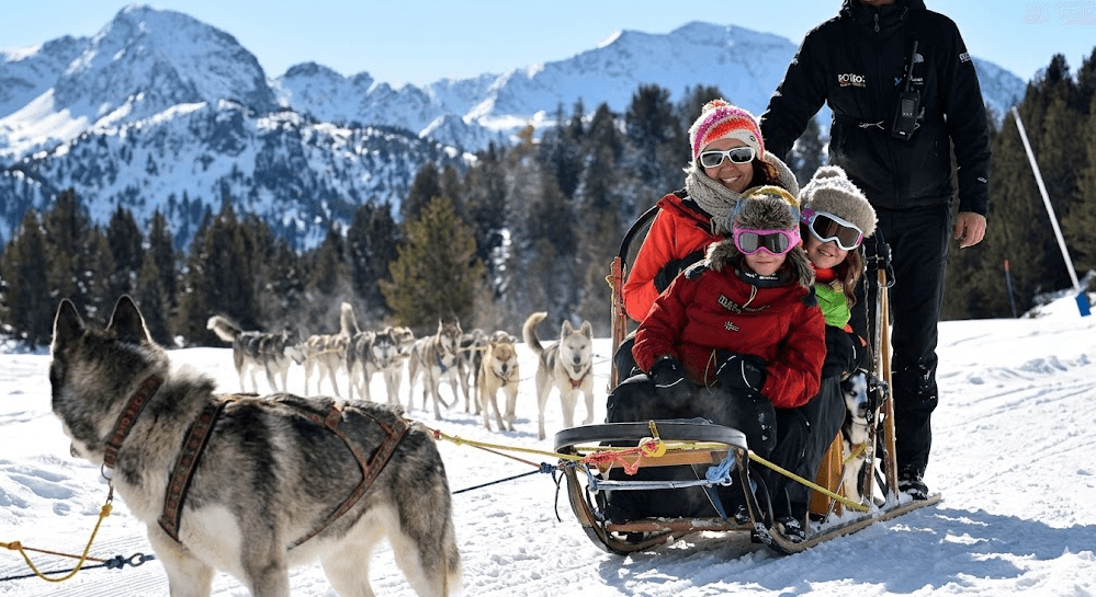 Una familia, compuesta por una madre y dos niños, pasea en un trineo de perros tirado por un equipo de huskies en un paisaje montañoso nevado. Un musher se encuentra detrás del trineo, guiando a los perros.