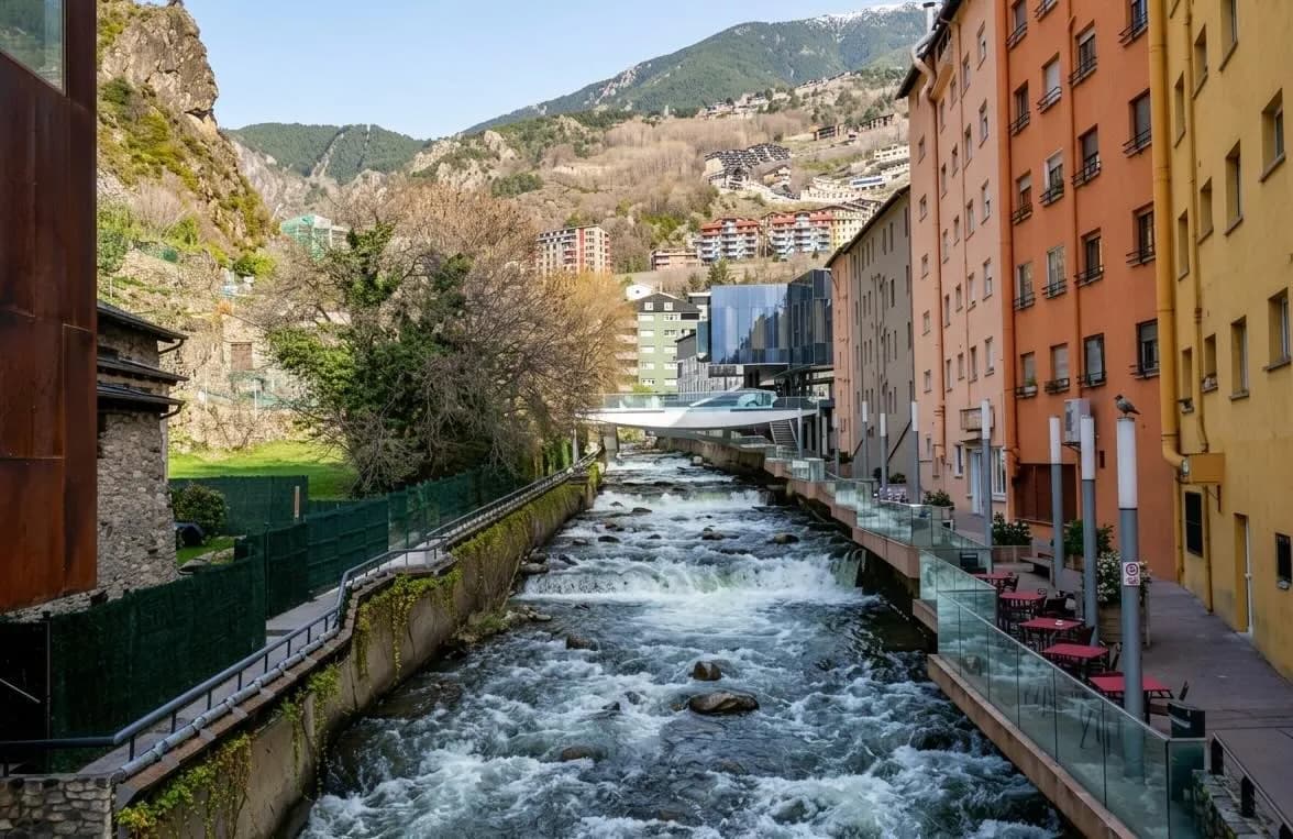 Un riu amb ràpids flueix a través d'una ciutat, flanquejat per edificis de colors i un pont modern, amb muntanyes al fons.
