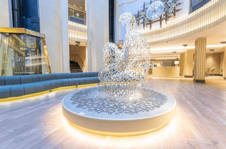 Elegant vestíbul d'hotel que presenta una gran escultura central d'una figura humana asseguda, construïda íntegrament amb lletres metàl·liques blanques. L'entorn compta amb terres de fusta clara, un sofà corbat blau, il·luminació càlida integrada i un gran rellotge decoratiu d'engranatges a la paret superior