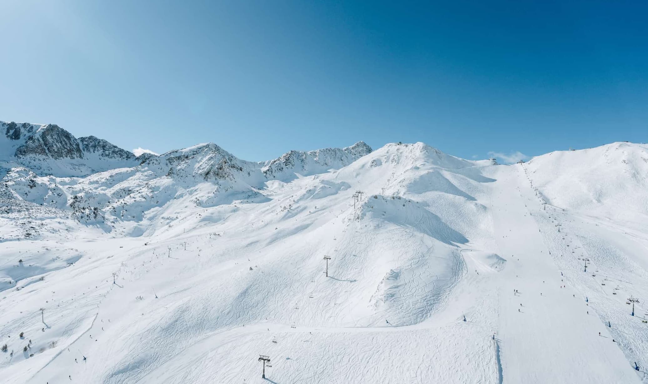 Vista aérea de una vasta estación de esquí montañosa cubierta de nieve bajo un cielo azul claro. Numerosos telesillas ascienden por las pistas, que están marcadas con huellas de esquí y salpicadas de pequeñas figuras de esquiadores.
