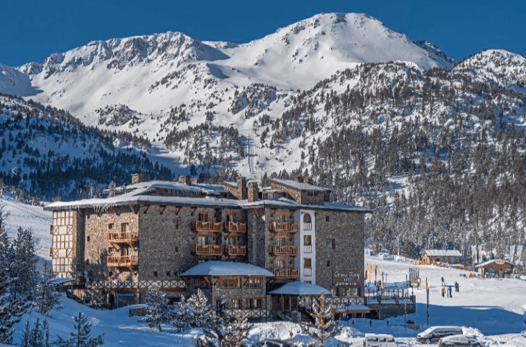 Un gran hotel de piedra y madera con balcones se alza en un paisaje montañoso nevado bajo un cielo azul claro. Pistas de esquí y pinos cubiertos de nieve rodean el edificio, con picos nevados distantes al fondo.