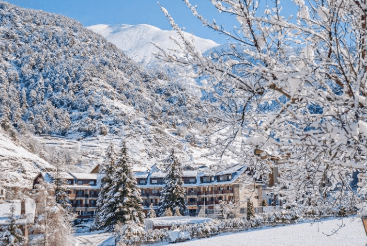 Un edificio de hotel de varias plantas con tejados rojos está rodeado de pinos y montañas cubiertas de nieve bajo un cielo azul brillante. Todo el paisaje está cubierto de nieve blanca y fresca.