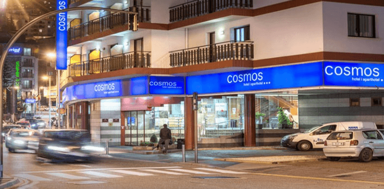 Vista nocturna de la fachada del Hotel & Aparthotel Cosmos en una esquina urbana, destacando sus grandes letreros luminosos de color azul. Se observa la entrada principal acristalada, balcones con barandillas de madera y el movimiento de coches en la calle bajo la luz de las farolas.