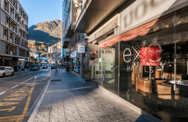 Vista de una calle urbana soleada en una zona de montaña, con la entrada acristalada del hotel Exe en primer plano a la derecha. Se observa una acera amplia con peatones, una carretera con tráfico moderado y una gran montaña escarpada al fondo bajo un cielo azul despejado