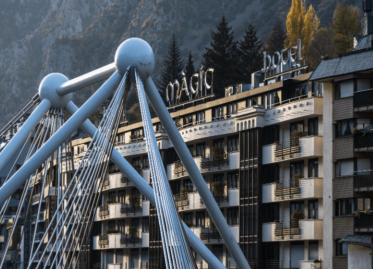 Fotografia en angle contrapicat del Pont de París a Andorra la Vella, mostrant la seva estructura de tirants d'acer i esferes metàl·liques en primer pla. Al fons, s'aprecia la façana de l'Hotel Màgic Andorra amb el seu rètol lluminós, envoltat de vegetació de muntanya i edificis residencials sota una llum de capvespre.