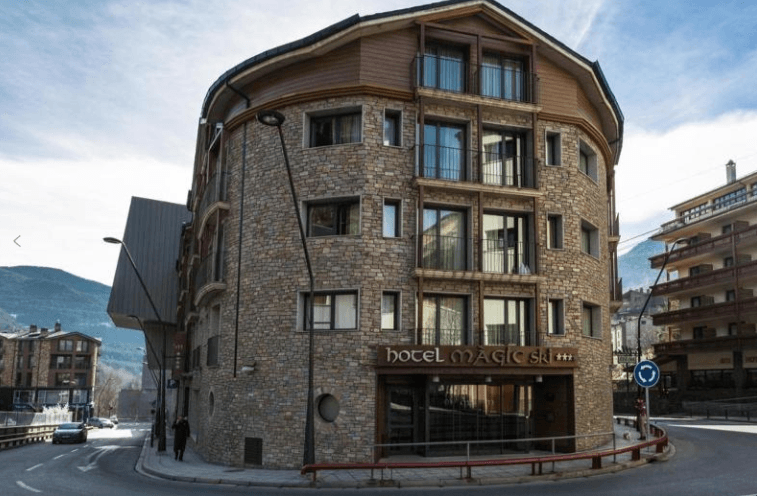 Un edificio de hotel de varias plantas con una fachada curva de piedra y balcones de madera se encuentra en una esquina. El letrero "Hotel Magic Ski" es visible en el edificio, con montañas al fondo.