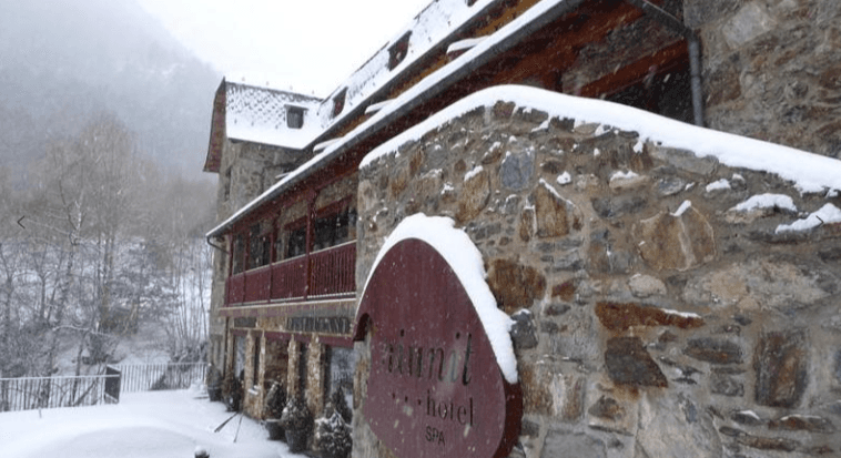 Un edificio rústico de hotel de piedra está cubierto de nieve fresca, con copos de nieve cayendo. Un letrero parcialmente visible dice "Niunit hotel SPA" junto a una pared nevada, con árboles y montañas cubiertos de nieve al fondo.