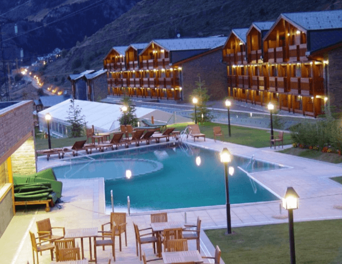 Piscina exterior iluminada al atardecer frente a un conjunto de edificios de estilo alpino en una zona de montaña, con tumbonas y mesas de madera alrededor y un ambiente tranquilo de hotel o resort.