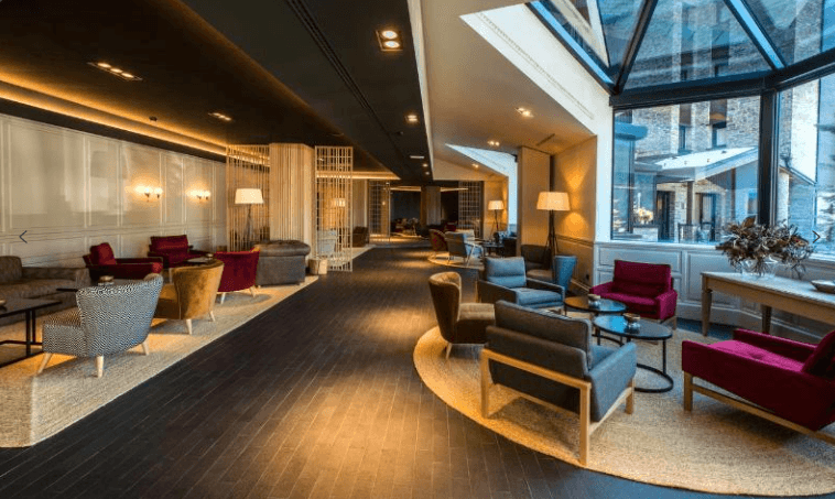 Un amplio y moderno salón de hotel con múltiples zonas de asientos que incluyen sillones y sofás coloridos, suelos de madera oscura y una iluminación cálida, además de una gran sección de ventanas con vistas al exterior de un edificio.