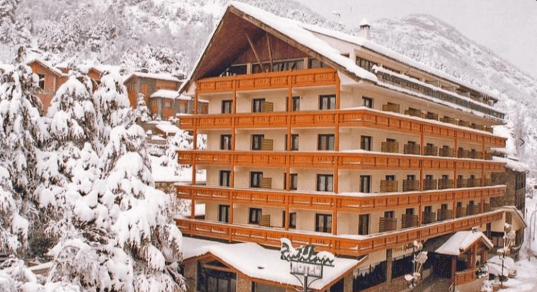 Un gran hotel de varias plantas con balcones de madera y tejado cubierto de nieve se alza entre pinos nevados y montañas en un paisaje invernal.
