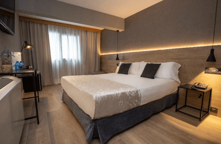 Habitación de hotel de diseño moderno con una cama doble vestida con ropa blanca y un camino de cama plateado. Destaca un cabecero de madera con iluminación LED cálida integrada, lámparas colgantes minimalistas de color negro, un escritorio lateral y suelos de tarima clara en un ambiente acogedor.