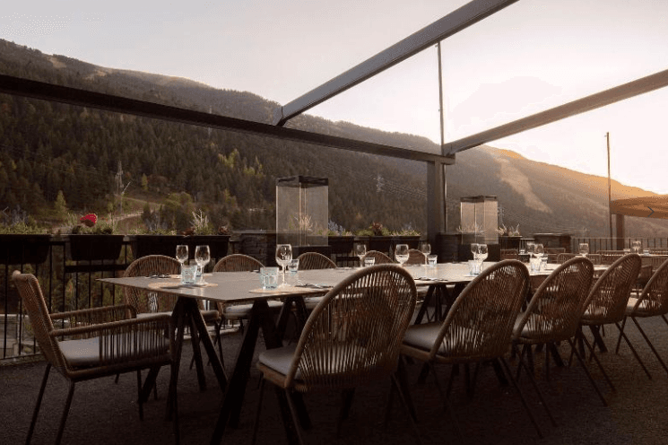 Vista de una terraza de restaurante de lujo al aire libre durante el atardecer, con mesas largas montadas con cristalería y sillas de mimbre de diseño. Al fondo se aprecia una panorámica de montañas frondosas con una pista de esquí visible y estufas de exterior de cristal, bajo una luz cálida de ocaso.