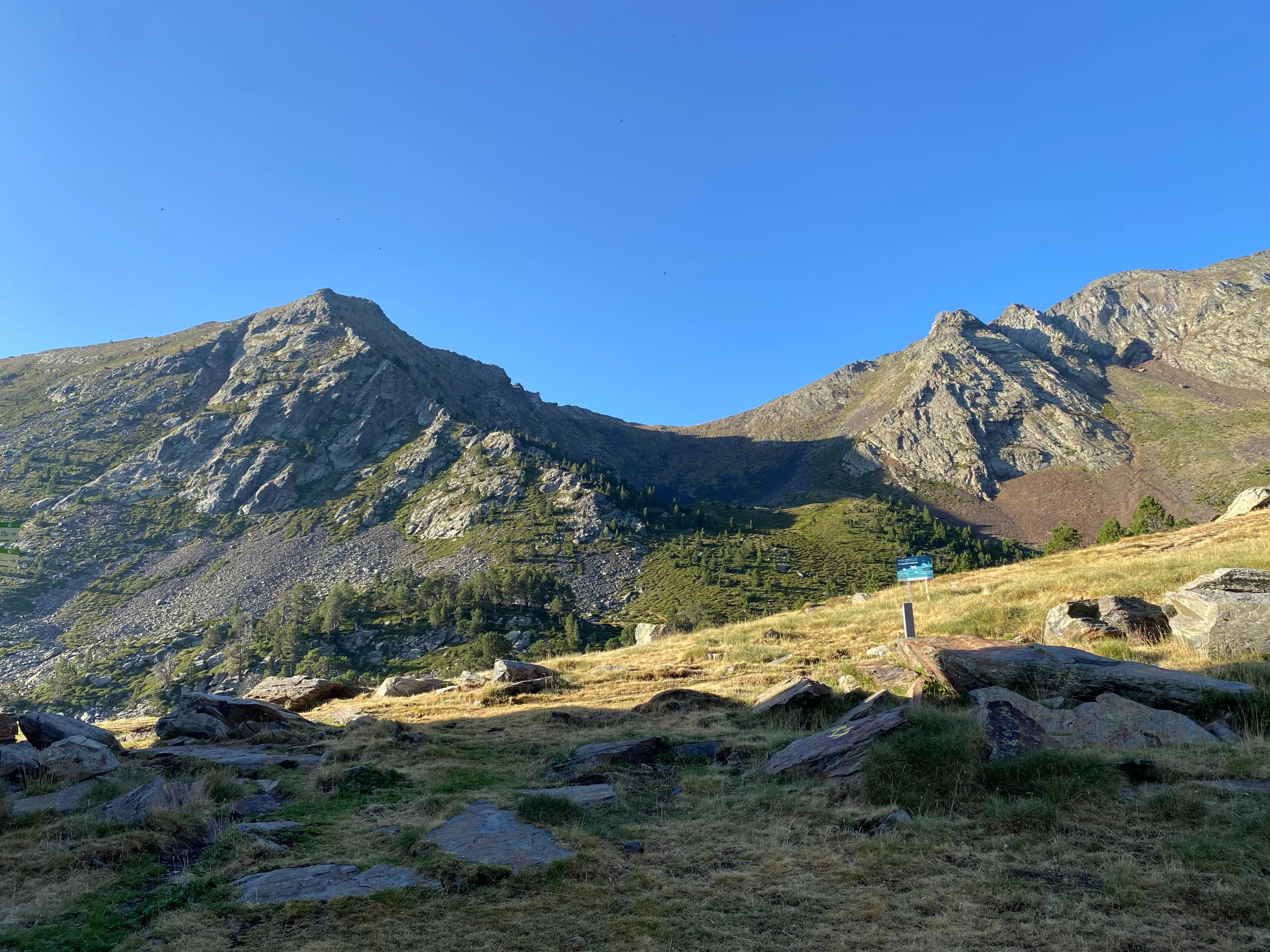 Una panoràmica d'un paisatge muntanyós amb dos cims rocosos sota un cel blau clar. El primer pla presenta herba seca i grans roques, que condueixen a vessants coberts de vegetació verda i arbres dispersos, amb un petit rètol blau visible a la dreta.