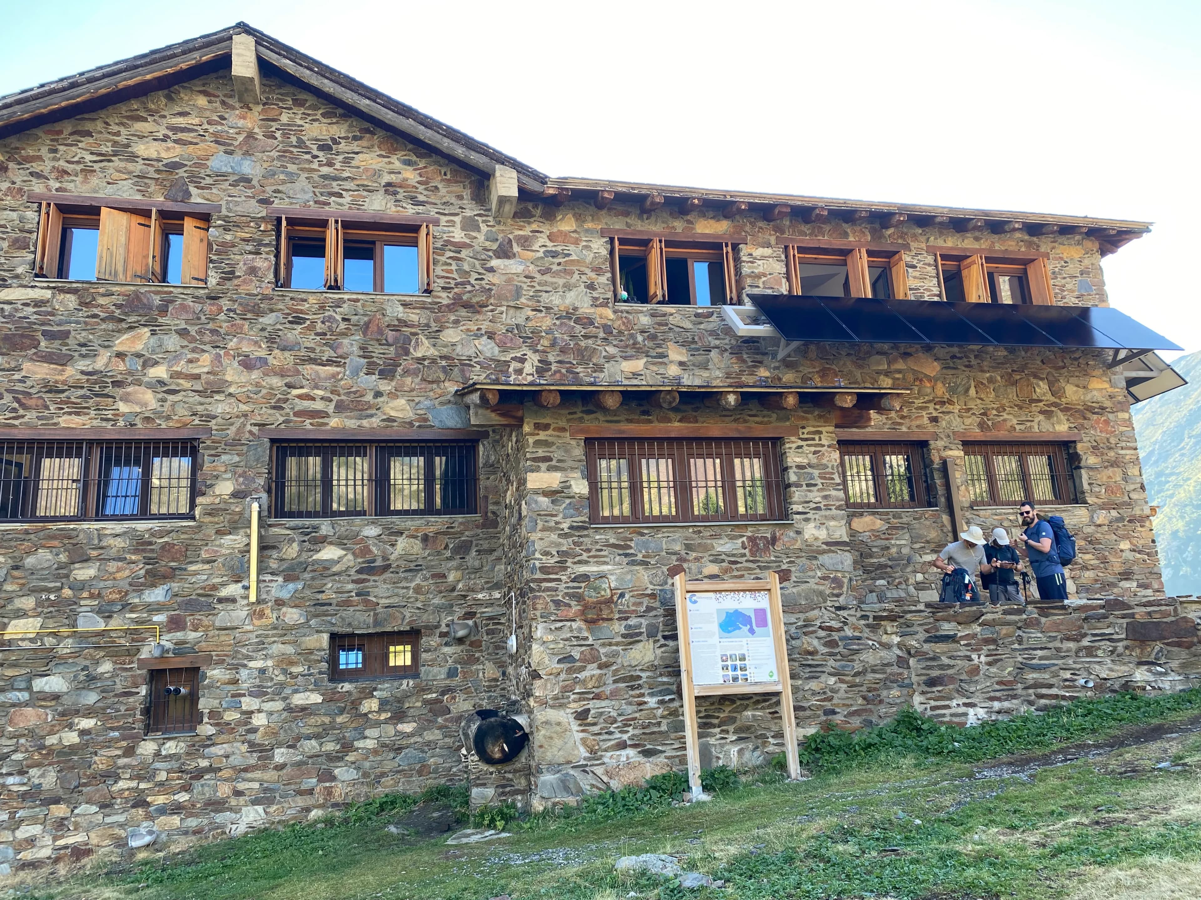 Un edifici de pedra de diversos pisos amb nombroses finestres, algunes amb persianes de fusta i altres amb reixes de metall, i panells solars a la façana superior. Tres excursionistes amb motxilles estan dempeus en un mur de pedra davant de l'edifici, amb un cartell informatiu de fusta a prop.