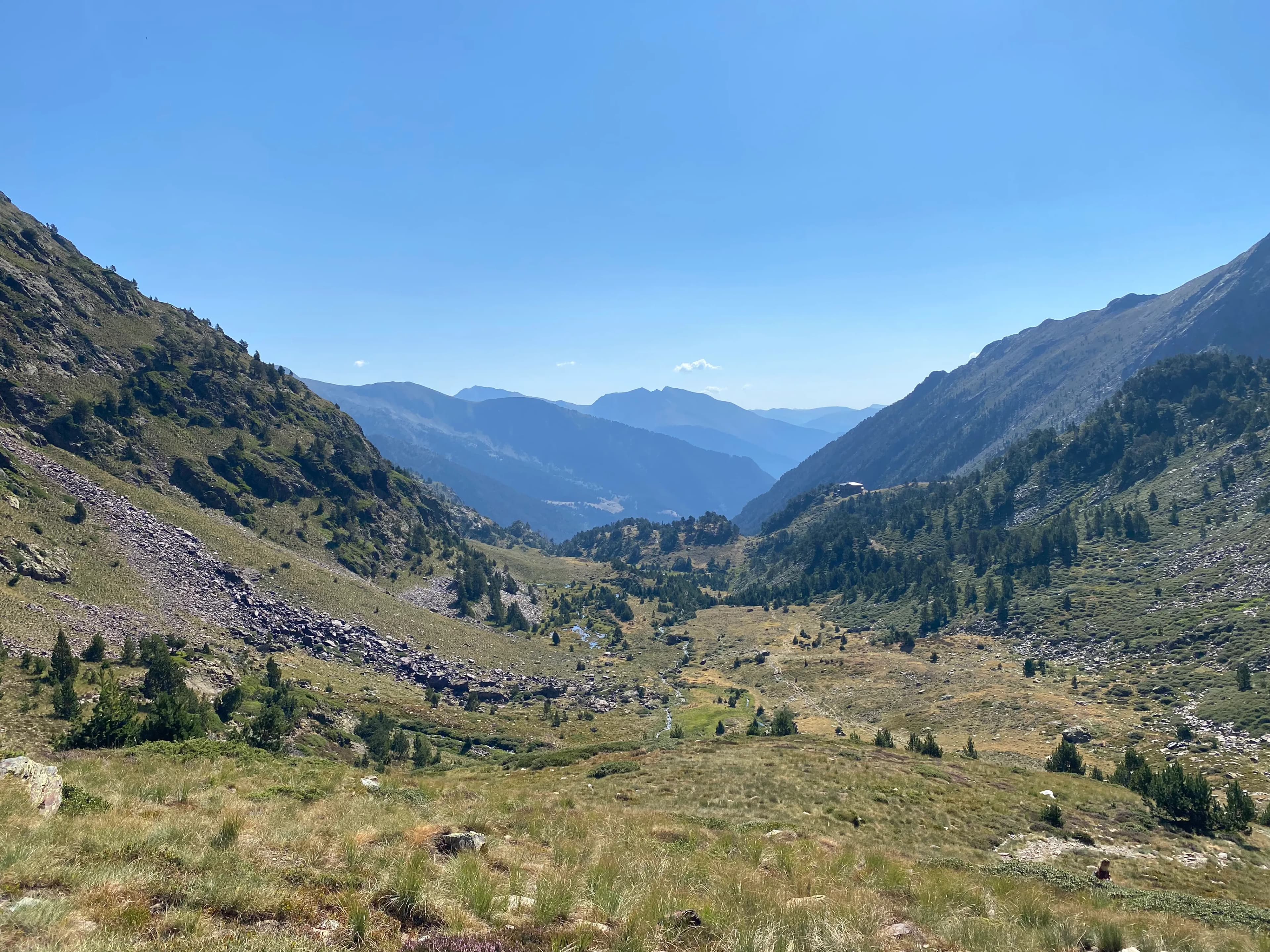 Una àmplia vall de muntanya amb vessants verds i rocosos, un rierol serpentejant i muntanyes boiroses a la distància sota un cel blau clar. Un petit edifici es troba en un vessant.
