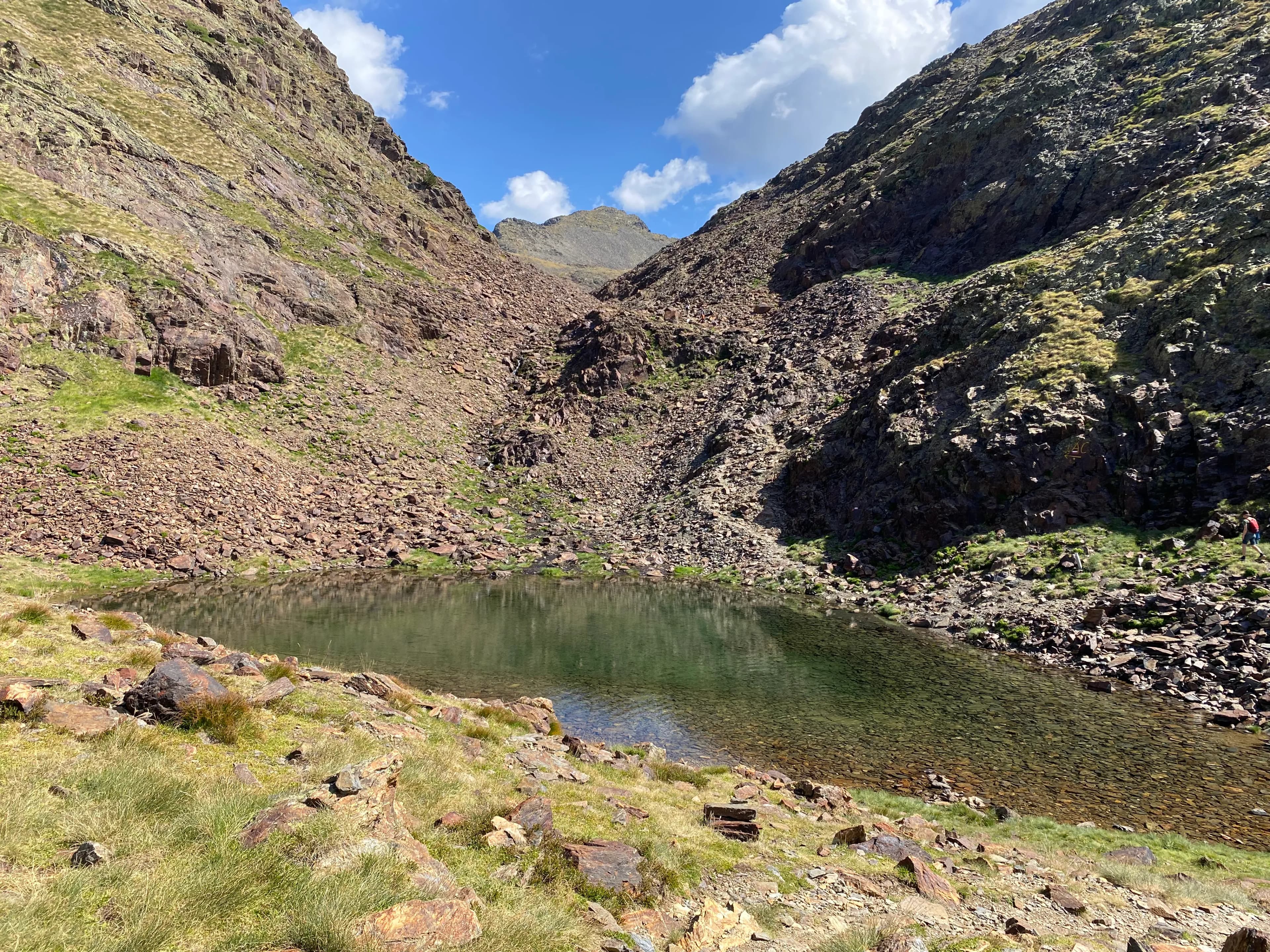 Un llac de muntanya clar encaixat entre vessants rocosos i escarpats coberts d'herba verda escassa sota un cel blau amb núvols blancs. Una persona es troba a la riba dreta.