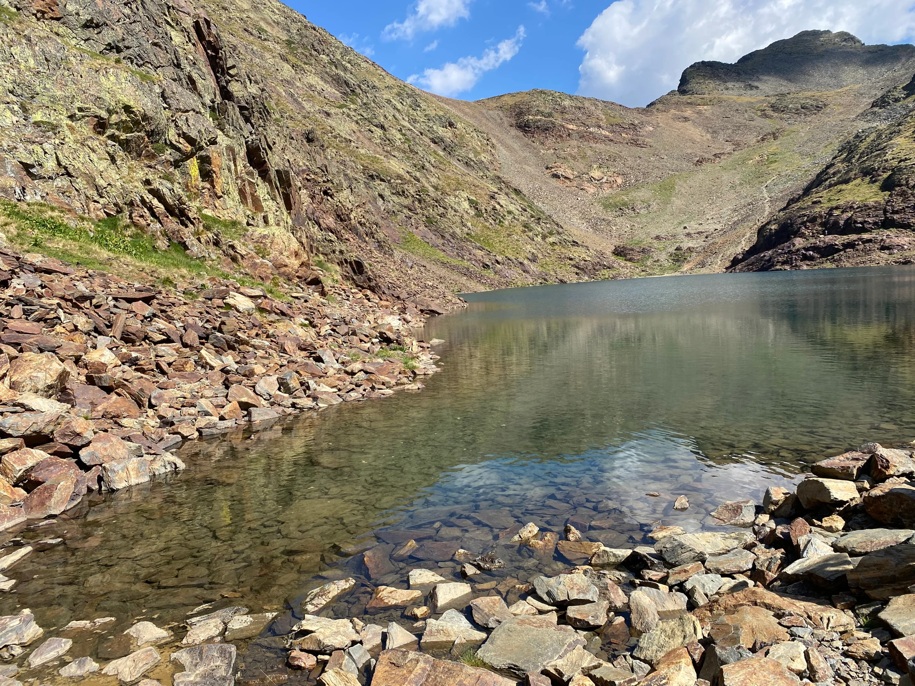 Un llac de muntanya transparent amb una riba rocosa i muntanyes escarpades sota un cel blau amb núvols blancs. L'aigua és transparent, revelant roques al fons del llac prop de la vora.