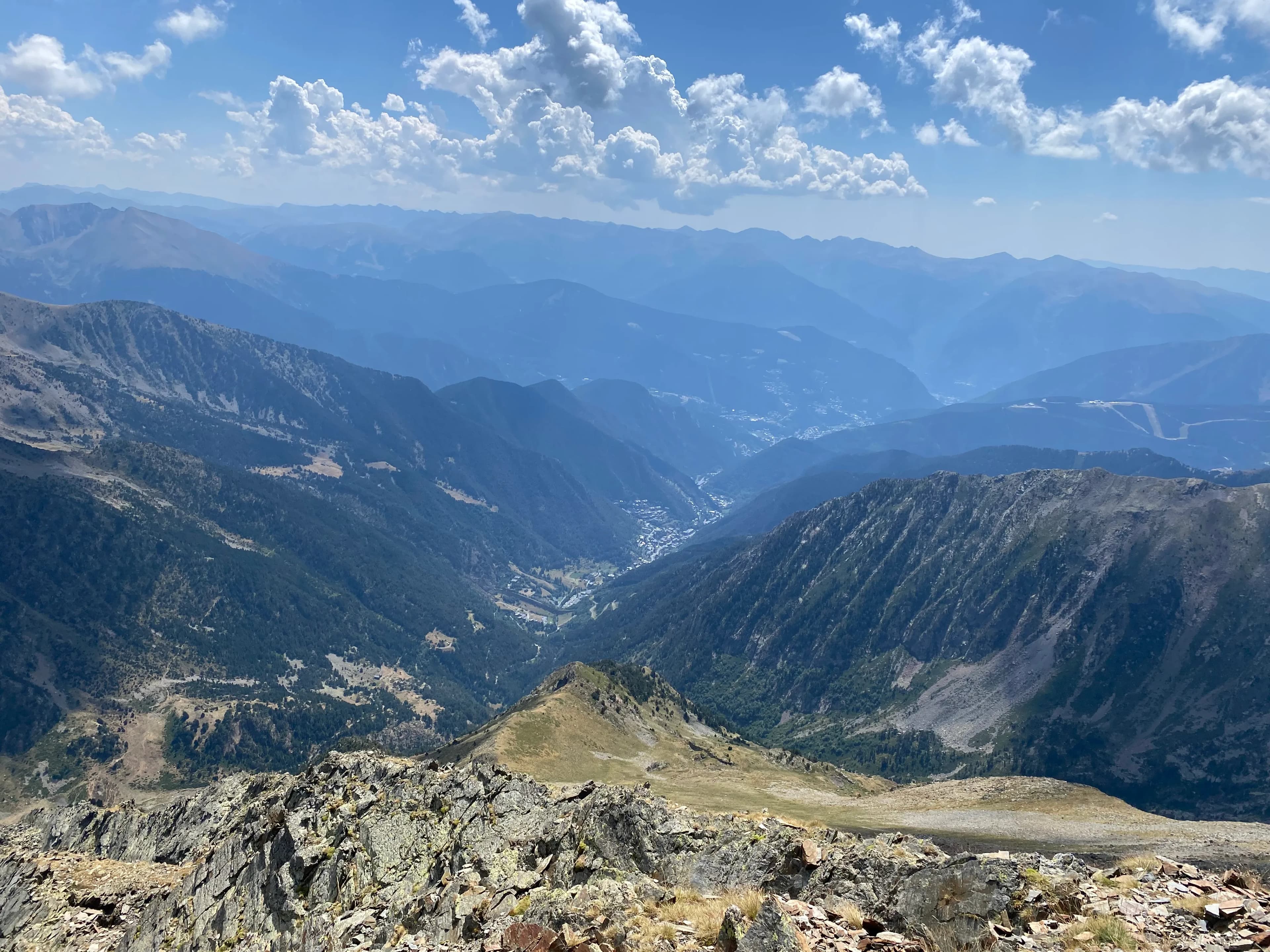 Una vista panoràmica d'una vasta serralada amb una vall i un petit poble encaixat entre els cims. El primer pla rocós dóna pas a vessants boscosos i muntanyes blaves boiroses distants sota un cel parcialment ennuvolat.