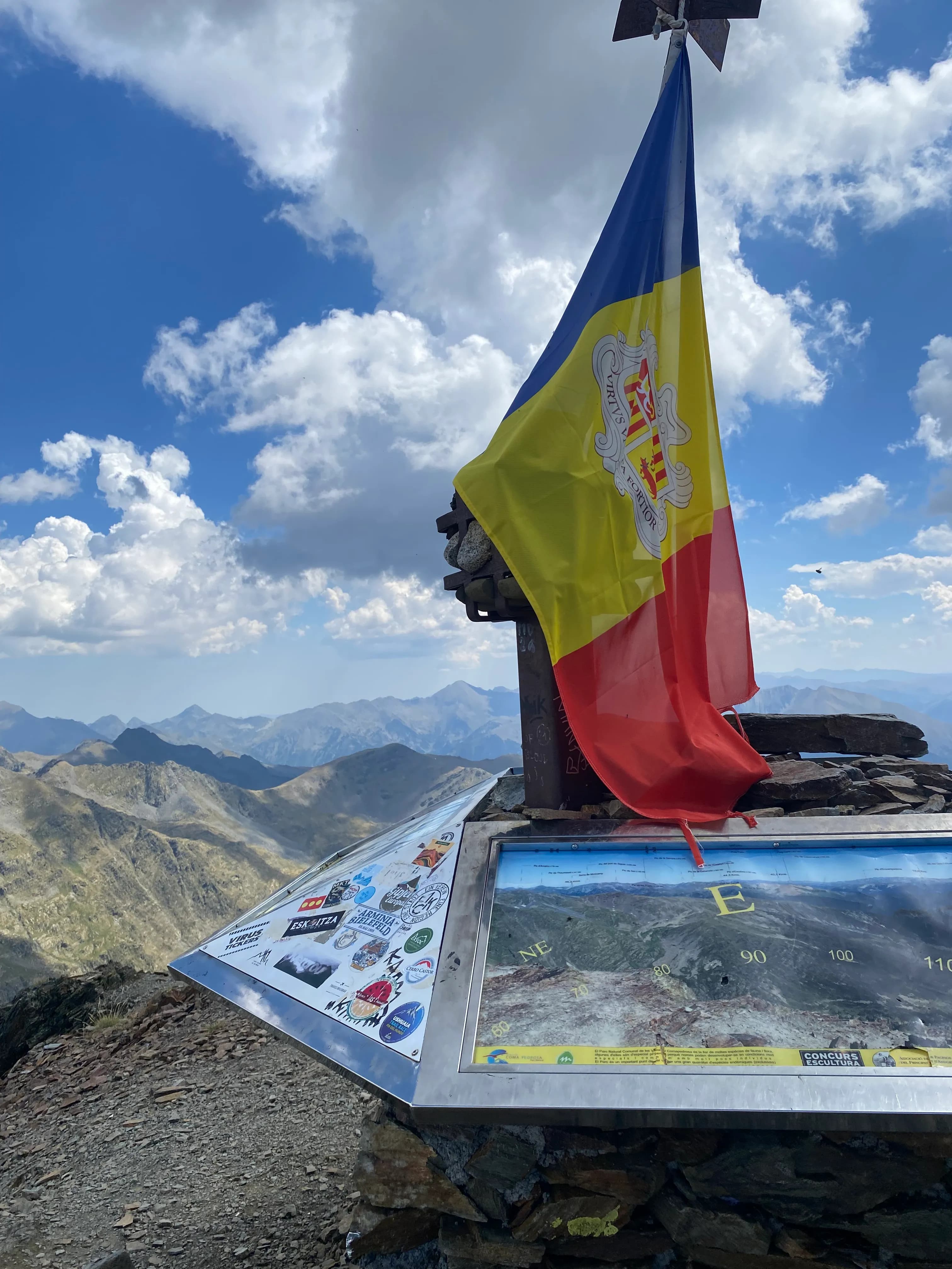 La bandera d'Andorra oneja des d'un marcador de cim en una muntanya, amb un panell informatiu que mostra un mapa i adhesius, i una vista panoràmica de muntanyes sota un cel blau ennuvolat.
